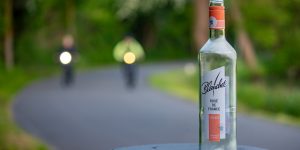 Bild einer Weinfalasche auf einem Mülleimer am Rande einer Straße. Im Hintergrund sind 2 Fahrradfahrer erkennbar.